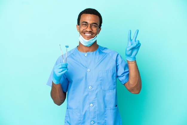 Dentista afro-americano segurando ferramentas sobre fundo azul isolado, sorrindo e mostrando sinal de vitória