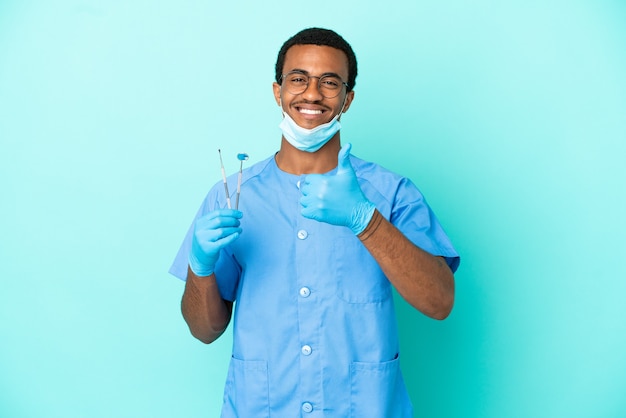 Dentista afro-americano segurando ferramentas sobre um fundo azul isolado e fazendo um gesto de polegar para cima