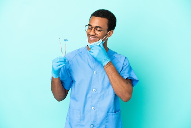 Dentista afroamericano sosteniendo herramientas sobre fondo azul aislado mirando hacia el lado y sonriendo