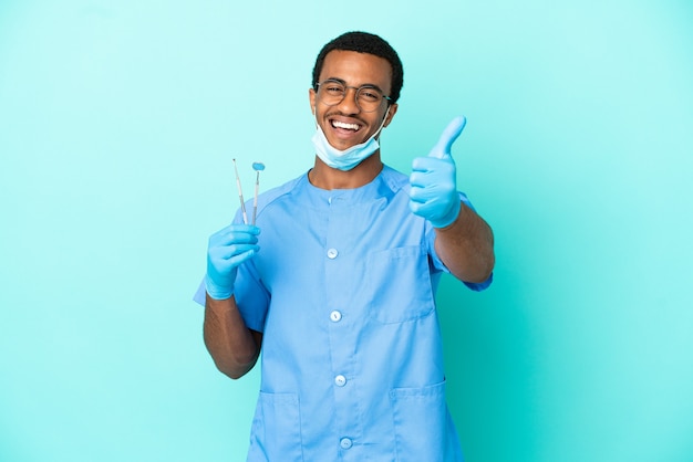 Dentista afroamericano sosteniendo herramientas sobre fondo azul aislado con los pulgares hacia arriba porque algo bueno ha sucedido