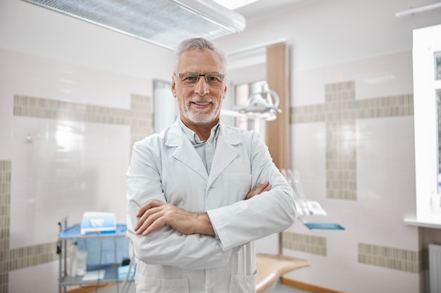 Dentista anciano alegre vistiendo una bata de laboratorio con los brazos cruzados sobre el pecho mientras sonríe a la cámara