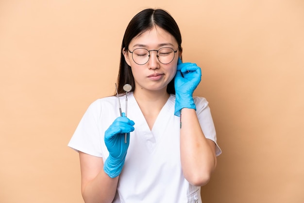 Dentista chinesa segurando ferramentas isoladas em fundo bege frustrada e cobrindo os ouvidos