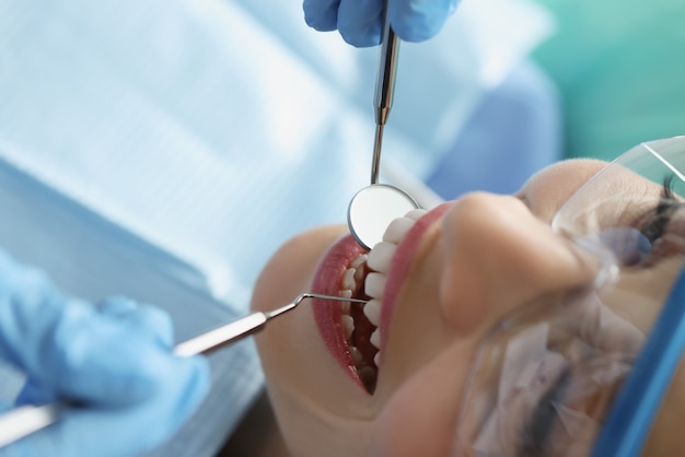 Dentista com luvas de proteção, examinando os dentes de uma paciente do sexo feminino usando instrumentos de metal na clínica