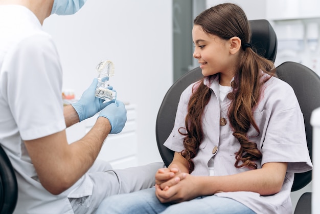 El dentista se comunica con su paciente, muestra en la mandíbula cómo cepillarse los dientes correctamente. La niña es una adolescente en un sillón dental.