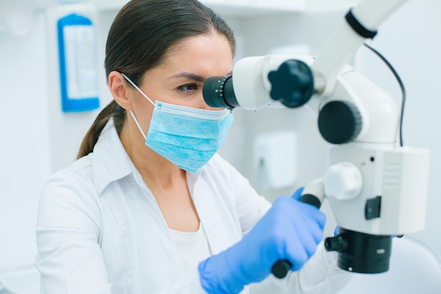 Dentista confiante com luvas de borracha e máscara médica, tocando as alças de um microscópio e olhando pelas oculares