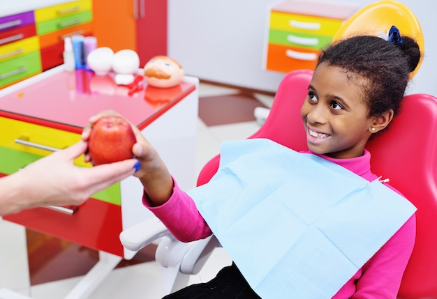 El dentista le da una manzana roja madura a una niña afroamericana negra en una silla dental.