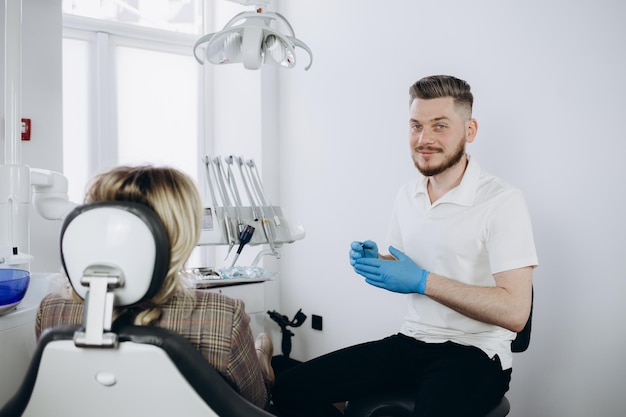 Dentista de homem com máscara facial e óculos fazendo tratamento para paciente loira segurando instrumentos odontológicos usando luvas de borracha Estomatologia odontologia moderna clínica odontológica conceito