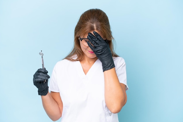 Dentista de meia-idade segurando ferramentas sobre fundo azul isolado com expressão cansada e doente