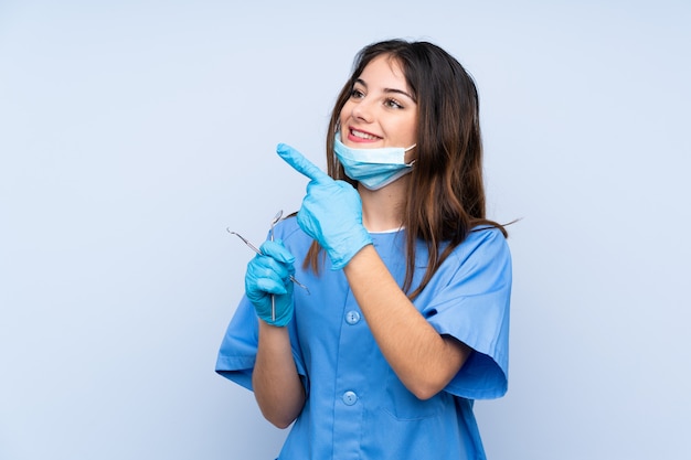 Dentista de mulher segurando ferramentas sobre parede azul, apontando com o dedo indicador