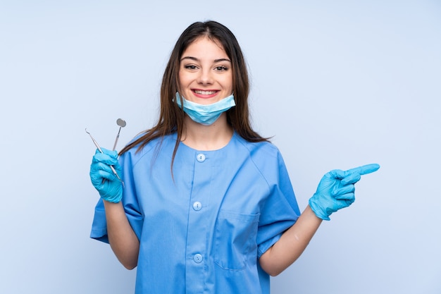Dentista de mulher segurando ferramentas sobre parede azul, apontando o dedo para o lado
