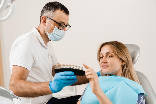 Dentista demonstra guia de tons de cor dos dentes para clareamento dental para paciente mulher na clínica odontológica odontologia mulher olhando para folheados ou implantes amostras de correspondência de cores dos dentes nas mãos do médico