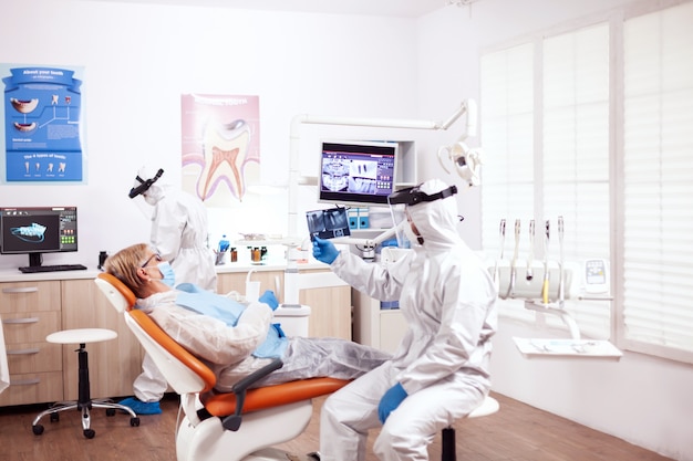 Dentista em equipamento de proteção agasint coroanvirus segurando o raio-x do paciente sentado na cadeira. mulher idosa com uniforme de proteção durante o exame médico na clínica odontológica.