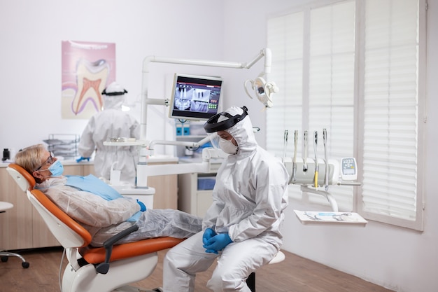Dentista em equipamento de proteção agasint coroanvirus segurando o raio-x do paciente sentado na cadeira. mulher idosa com uniforme de proteção durante o exame médico na clínica odontológica.