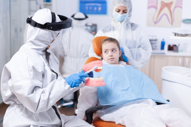 Dentista enseñando a niño a cepillarse los dientes vestidos con ppe. Niña en traje de ppe durante covid19 y madre escuchando estomatólogo hablando de higiene dental en clínica de dentista con modelo de mandíbula.