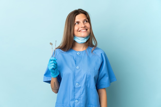 Dentista eslovaco sosteniendo herramientas aisladas sobre pared azul pensando en una idea mientras mira hacia arriba