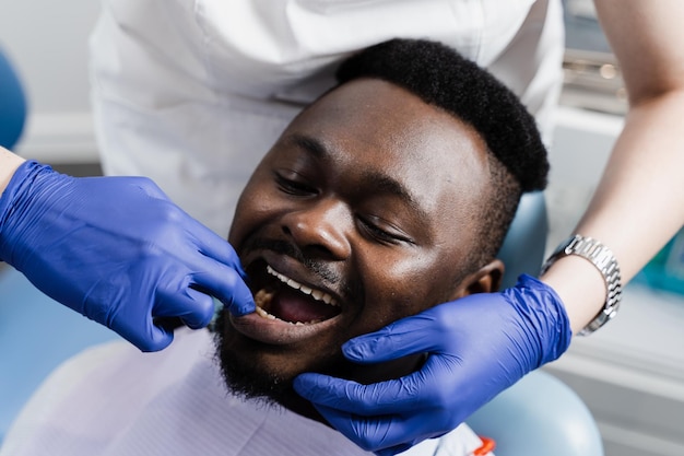 El dentista examina los dientes y la boca del hombre africano con las manos enguantadas azules El dentista toca los dientes del paciente con las manos en odontología