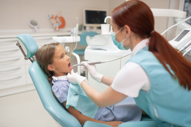 Dentista examinando. Dentista pelirrojo con guantes blancos examinando colegiala en la mañana