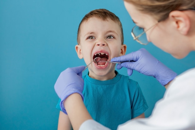 Dentista examinar los dientes del niño en la pared azul. Un pequeño paciente en el sillón dental sonríe. Dantist trata los dientes. Vista cercana del dentista que trata los dientes del niño pequeño en el consultorio del dentista.