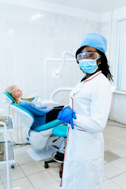 Dentista fêmea afro-americano de sorriso dos jovens na frente do paciente na clínica. conceito de clínica dentária.