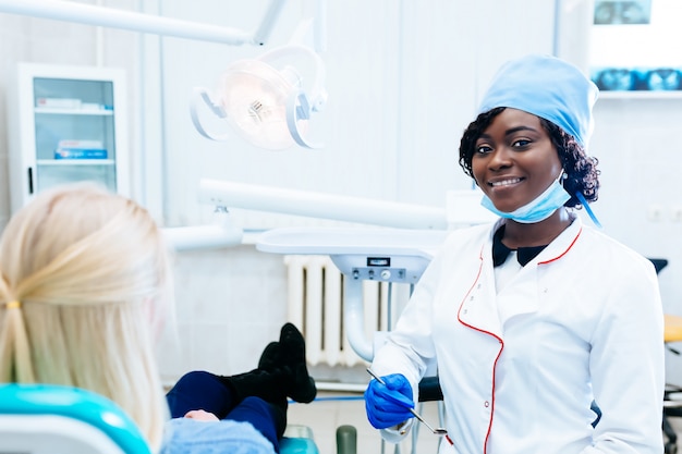 Dentista fêmea afro-americano que trata o paciente na clínica. Conceito de clínica dentária.