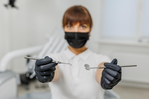 Dentista feminina com máscara facial preta uniforme branca e luvas mostrando ferramentas de estomatologia entre modernas