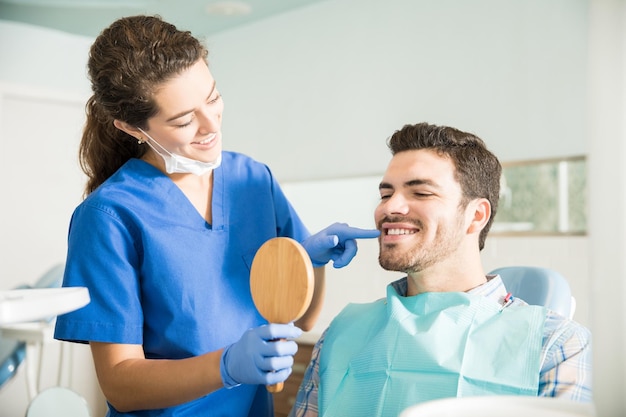 Dentista feminina sorridente mostrando espelho para homem adulto médio após tratamento na clínica odontológica