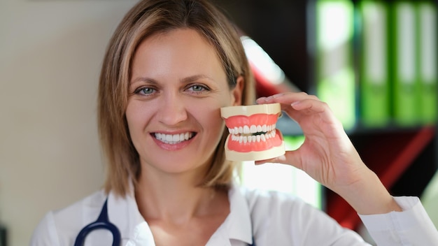 Dentista feminina sorrindo e mostra mandíbulas artificiais em sua mão médica estomatologista