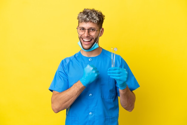 Dentista hombre caucásico sosteniendo herramientas aisladas sobre fondo amarillo celebrando una victoria