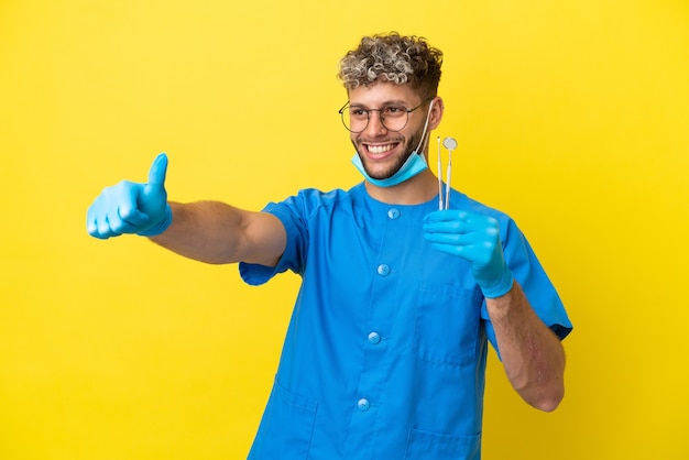 Dentista hombre caucásico sosteniendo herramientas aisladas sobre fondo amarillo dando un pulgar hacia arriba gesto