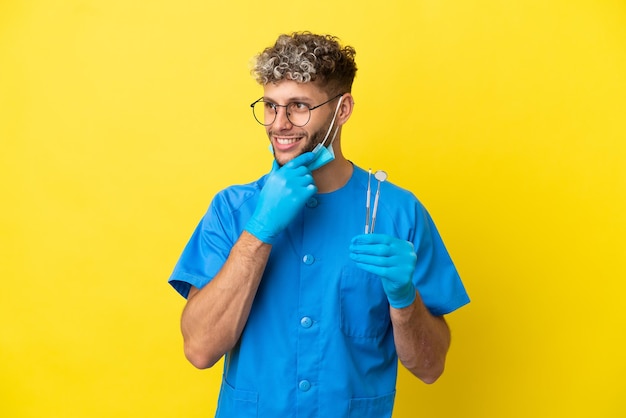 Dentista hombre caucásico sosteniendo herramientas aisladas sobre fondo amarillo mirando hacia el lado y sonriendo