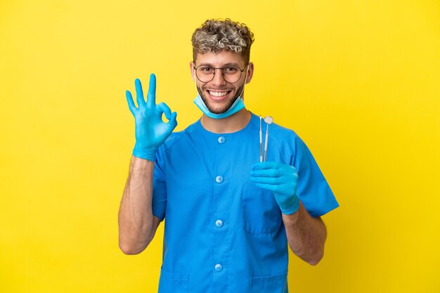 Dentista hombre caucásico sosteniendo herramientas aisladas sobre fondo amarillo mostrando signo ok con los dedos