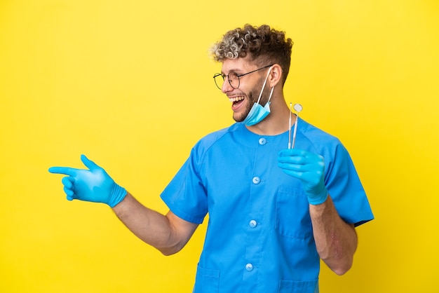 Dentista hombre caucásico sosteniendo herramientas aisladas sobre fondo amarillo que señala el dedo hacia el lado y presenta un producto