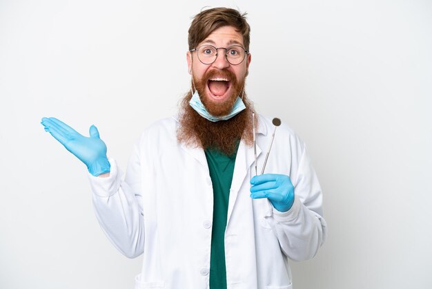 Dentista hombre rojizo sosteniendo herramientas aisladas sobre fondo blanco con expresión facial sorprendida