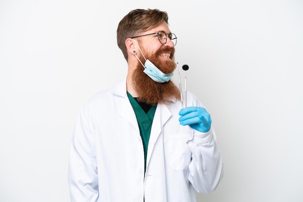 Dentista hombre rojizo sosteniendo herramientas aisladas sobre fondo blanco mirando hacia el lado