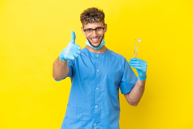 Dentista hombre rubio sosteniendo herramientas aisladas en el fondo con los pulgares hacia arriba porque algo bueno ha sucedido