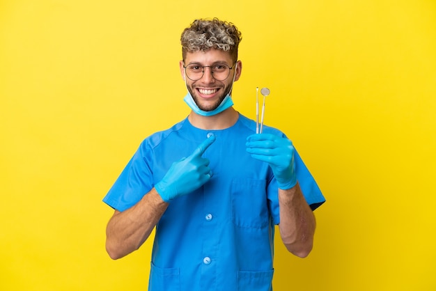 Dentista homem caucasiano segurando ferramentas isoladas em um fundo amarelo apontando para o lado para apresentar um produto