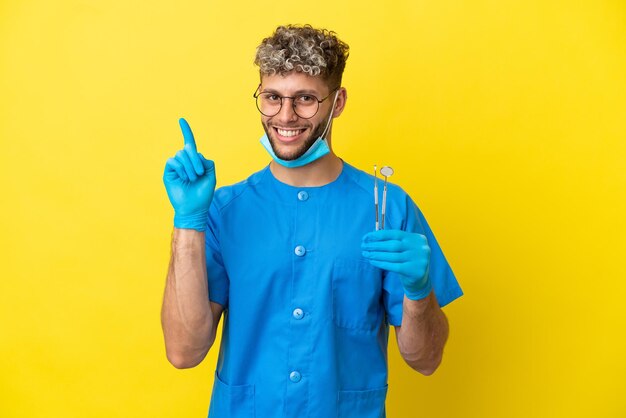 Dentista homem caucasiano segurando ferramentas isoladas em um fundo amarelo, mostrando e levantando um dedo em sinal dos melhores