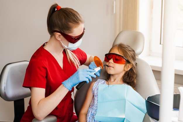 El dentista infantil trata y elimina la caries en un paciente Iluminación ultravioleta del procedimiento de llenado de dientes con fotopolímero Odontología infantil