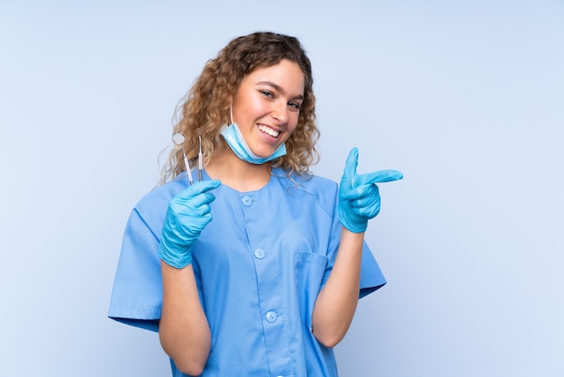 Dentista jovem loira segurando ferramentas isoladas em azul apontando o dedo para o lado