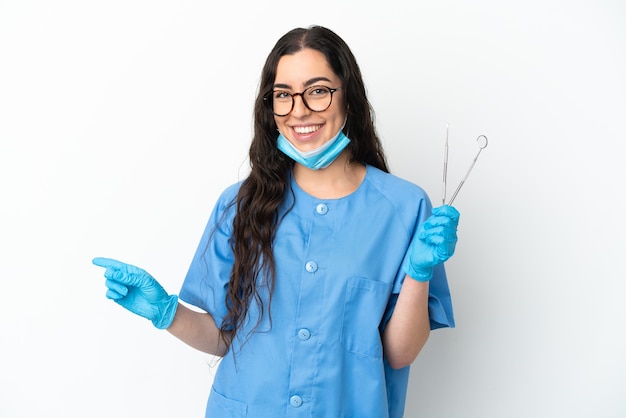 Foto dentista jovem segurando ferramentas isoladas na parede branca apontando o dedo para o lado
