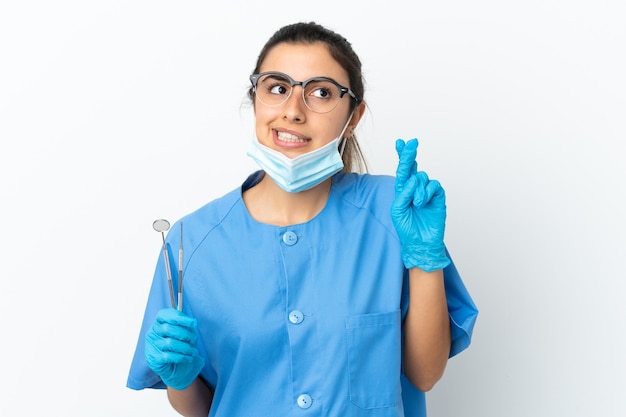 Dentista jovem segurando ferramentas isoladas no fundo branco, cruzando os dedos e desejando o melhor