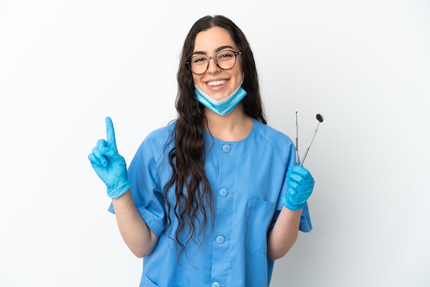 Foto dentista jovem segurando ferramentas isoladas no fundo branco, mostrando e levantando um dedo em sinal dos melhores