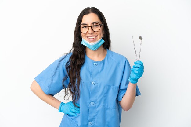 Foto dentista jovem segurando ferramentas isoladas no fundo branco, posando com os braços na cintura e sorrindo