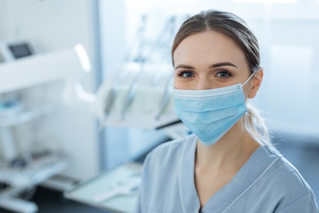 Foto dentista joven alegre posando en su oficina y sonriendo mientras usa una mascarilla