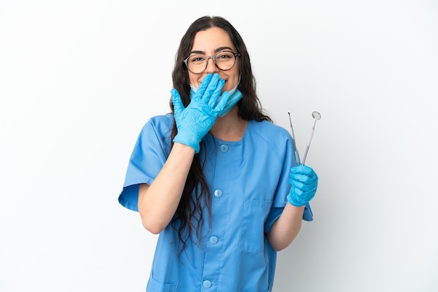 Dentista joven sosteniendo herramientas aisladas sobre fondo blanco feliz y sonriente cubriendo la boca con la mano