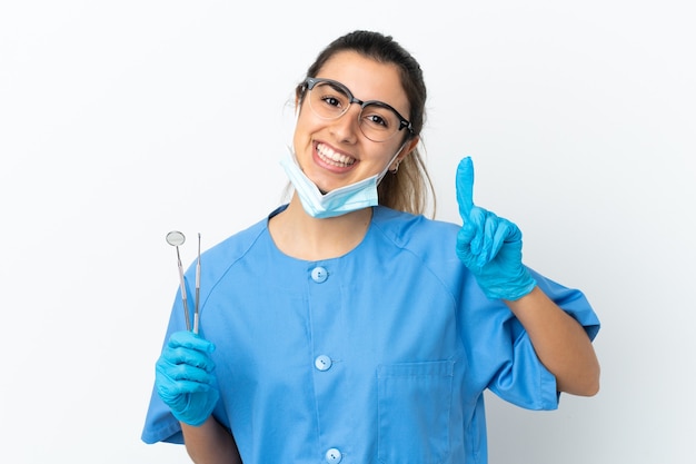 Dentista joven sosteniendo herramientas aisladas sobre fondo blanco mostrando y levantando un dedo en señal de lo mejor
