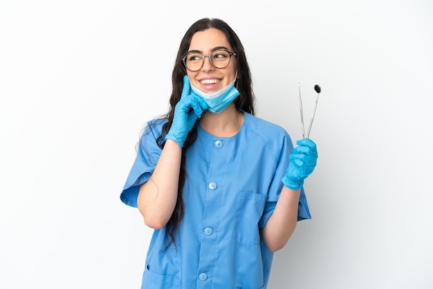 Dentista joven sosteniendo herramientas aisladas sobre fondo blanco pensando en una idea mientras mira hacia arriba