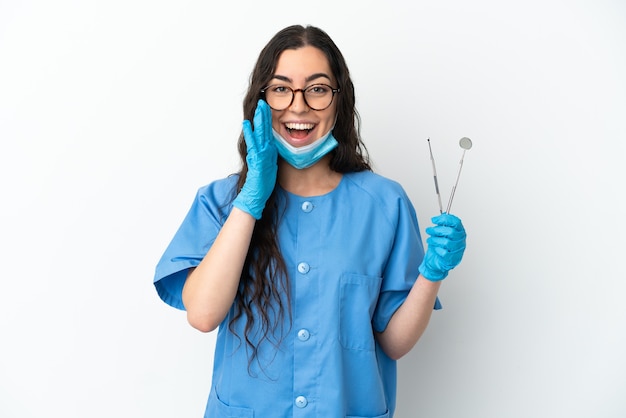 Dentista joven sosteniendo herramientas aisladas sobre pared blanca gritando con la boca abierta