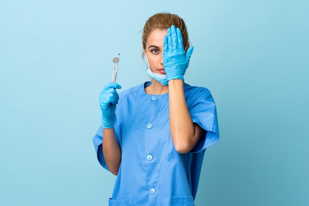 Dentista joven sosteniendo herramientas sobre azul aislado cubriendo un ojo con la mano