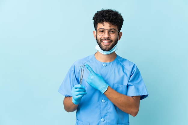 Dentista marroquí hombre sujetando herramientas aisladas en azul apuntando hacia el lado para presentar un producto